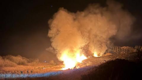 The scene of a large fire on woodland in Somerset. The orange blaze can be seen and there is smoke rising up to the sky. Small silhouettes of firefighters can be seen near the fire
