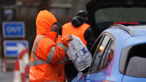 A woman in an orange jacket hands bottles of water through a car window.