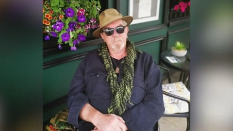 A man sitting outside. He has his hands crossed in front of him and his looking at the camera. He is wearing a hat, sunglasses, scarf, necklace and blue jacket. Behind him there is a table and chair and a building with a hanging basket of flowers on its wall.