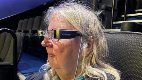An older lady with long grey hair sitting in a cinema with grey seats and blue carpet. She is wearing smart glasses over her usual glasses and is obviously looking at a screen out of shot. There is a grey wire coming from the side of the smart glasses.