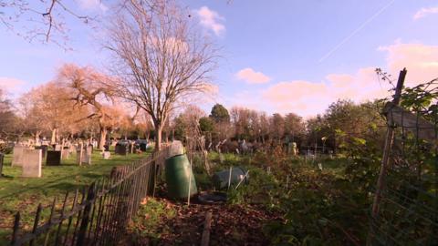 A green area to the left of a black fence shows graves stones. To the right, compost bins lean against the fence and on the ground and vegetation is growing in the background.