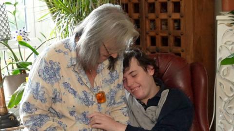 Chantal Chaervey rests on the arm of red leather chair which her son Harry sits in. She has shoulder length grey hair and wears a white blouse with blue flowers on it. Harry smiles as he rests his head on his mother's shoulder. He has dark hair and wears a grey and blue long sleeved top. 