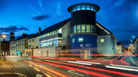 A long, curved street in Stornoway. A large arts centre is at the end of the road. The entrance is a tall blue and glass cylindrical building.