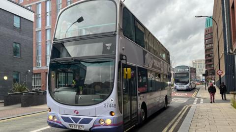 A First-branded purple double-decker bus travels along a road in Sheffield city centre, followed by another in the background.