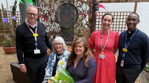 Two men and three women standing in front of the new garden. Two women are sitting down and smiling at the camera. One is holding a bouquet of flowers.
Either side of the women sitting down there are people standing. Everyone is smiling at the camera. 
