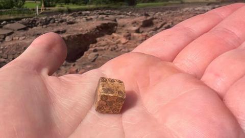 A person holds a small, light brown die with carved spiral shapes in the palm of their hand. 