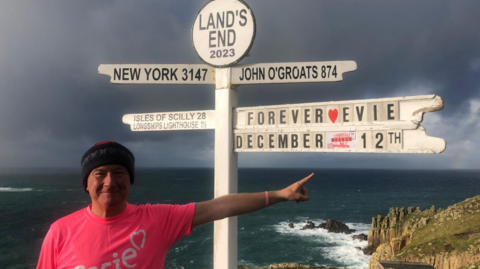 Dickie Mackness standing at a fingerpost at Land's End, with signs to John O'Groats and New York, and also one saying "Forever Evie, December 12th". He is wearing a pink t-shirt saying Evie and the sea is in the background with waves crashing over some rocks. 
