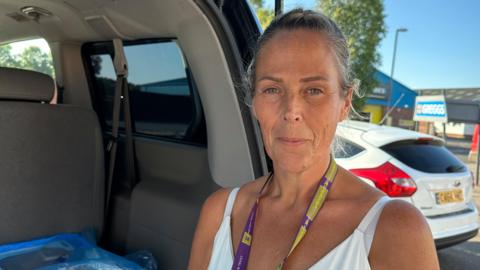 A woman in a white vest standing at the open boot of a van in a car park looking at the camera