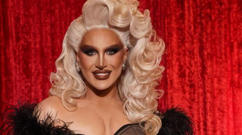 The Vivienne, who has curly blonde hair and is wearing a black off-the-shoulder dress, smiles at the camera in front of a red velvet curtain.