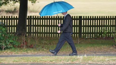 Man walking in rain holding a large blue golfing umbrella with fields behind