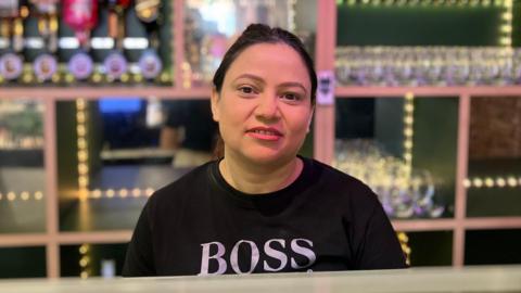 A woman in front of a bar with optics behind her. She wears a black T-shirt with a BOSS logo.