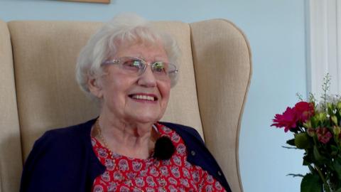 A picture of Beryl sat in a large cream coloured chair. She is wearing a red outfit with a navy blue cardigan. She is wearing glasses, while smiling.