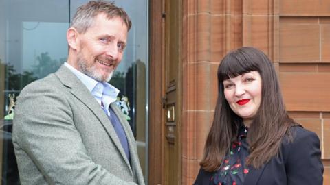 A man with short grey-brown hair and a greying beard in a grey jacket with a blue jumper and pale blue shirt stands next to a woman with long brown hair, bright red lipstick and a patterned blouse and black jacket on the steps of Dumfries and Galloway Council.