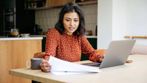 A woman looks over bills while sitting at a desk in an orange dress.