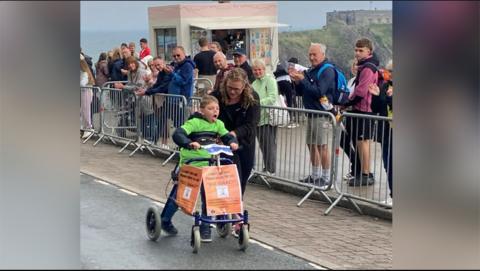 Boy with short fair hair wearing a green and blue shirt and blue trousers pushing a walker with orange posters fixed to it past a line of spectators behind metal rails.  The sea is visible in the background.