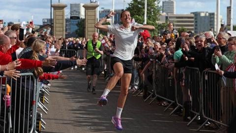 Former Lioness Jill Scott arrives at the Stadium of Light in Sunderland, as she finishes a five day cycle and marathon challenge for Sport Relief. She is leaping in the air as she runs through a channel between barriers, behind which are a crowd of people cheering and taking photographs. She has purple and pink trainers, black shorts and a white t-shirt. She is beaming.