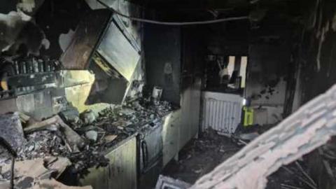 A kitchen that has been gutted by a fire. There is ash and rubble and all of the kitchen is black and burnt.