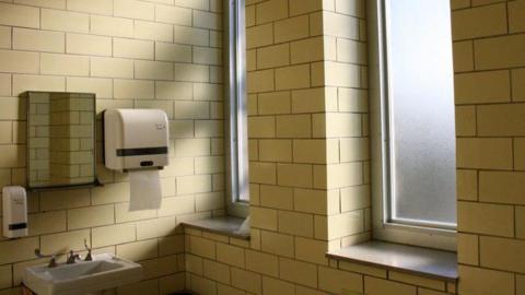 Photo of yellow-tiled restroom. There is a sink with a mirror, soap dispenser and paper dispenser above. There are two windows on the wall beside the sink. 