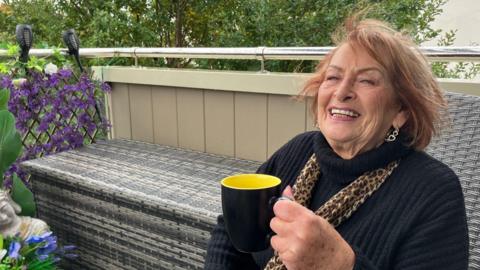 A smiling woman with a mug in her hands, sat in her chair on a balcony with purple foliage in the background