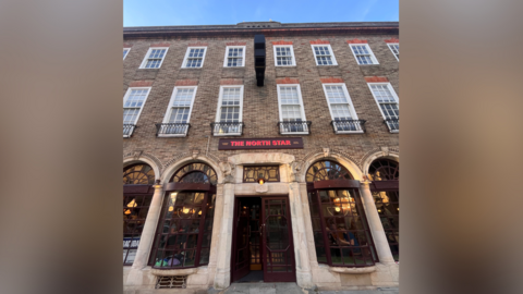 An exterior image of The North Star pub next to the Royal Pavilion in Brighton and Hove, East Sussex. The pub has brown doors and window frames, and has a sign above the door which says 'The North Star'. The sign is brown and the text within it is red.