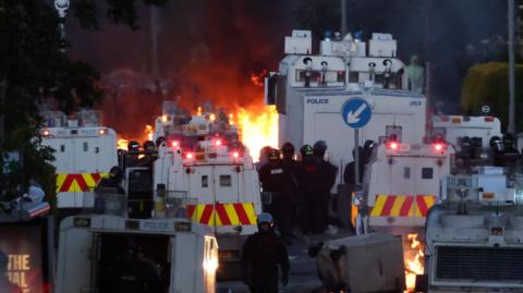 Police officers wearing riot gear stand beside vans as fires are lit in the distance.