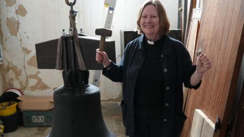 A woman vicar in black, wearing her dog collar, smiles at the camera. She has brown hair in a bob. She is holding a mallet in one hand and is standing next to a church bell 