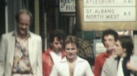 The five members of Landscape walking down a London street in the direction of the camera.  Their outfits are of the time and stylish.  The member on the left wears a floral shirt and white cream jacket, the man in the middle is a little shorter and appears to be wearing a white jumpsuit.  The member on the far right in the foreground has a grey jacket, black shirt and white tie on. The two members behind them are wearing a red jacket and a red shirt.  There is a road sign behind with listings for Aylesbury and St Albans and North West.