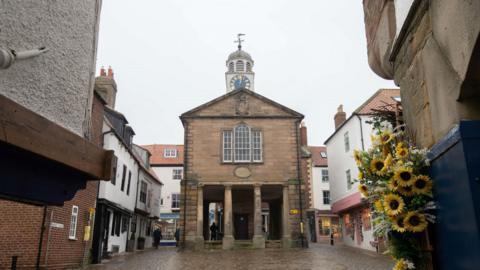 A grey stone building with a large arched window and columns supporting it at the front stands in market place with buildings on either side and behind. There is a blue clock on a white clock tower on top of the building. In the foreground on the right hand side is a bunch of flowers.