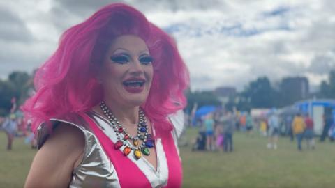 A drag queen with long cerise-pink hair and a silver and pink top. She is standing in a festival field full of people and stalls.