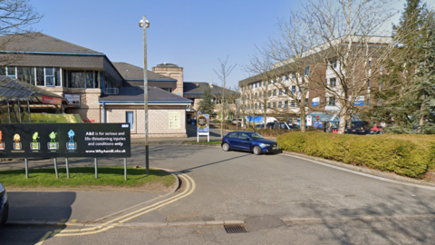 A street view of the Royal Lancaster Infirmary. It is made up of a number of of brick buildings with blue windows. An ambulance can be seen behind an A&E sign.