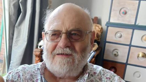 Ron Bailey, a man with a white beard and glasses, and a bald head. He is wearing a patterned open neck shirt and sitting next to a window in his home
