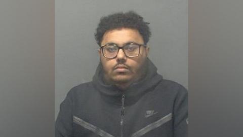 Police mugshot of Ahmed Omar wearing a dark-coloured hoodie and dark-rimmed glasses