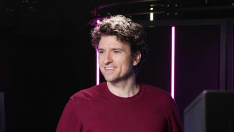 Greg James smiling left of the camera. He has curly hair and is wearing a maroon t-shirt. He's in a radio studio lit up with pink lights.