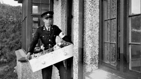 A black and white photo of a young man in a military-style uniform with a white armband on with a cross on it. He is at the door of a house, bringing in a drawer of medical equipment.