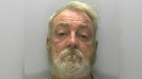 Mugshot of Edward Hanbury with white hair and a beard looking straight ahead. He is wearing a cream jumper and there is a grey background