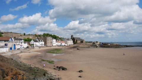 A coastal village overlooks a large beach with the tide out. There are blue skies and clouds above, with a few people walking along the sand. 