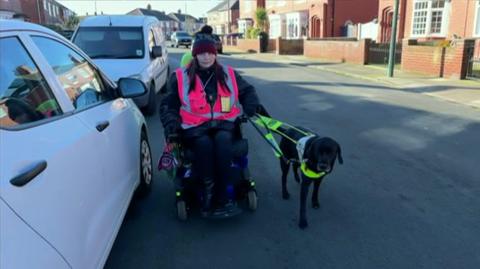 A woman sitting in an electric wheelchair in the middle of a road, with a guide dog beside her. Cars are parked to the left, with houses to either side. She is wearing a purple bobble hat, dark clothes and a pink hi-vis jacket. The dog is black and wears a bright green harness and lead.