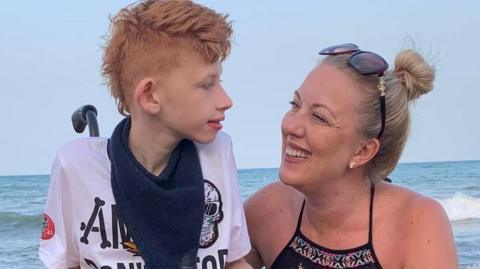 Mother Claire Stockton has blonde hair tied back and she is wearing a black summer dress. She is sat down smiling at her son who is a wheelchair user and is now 20 years old. Her son is disabled and has a rare condition called ReNU syndrome. He is wearing a white t-shirt with a blue scarf and is looking back at his Mum, Claire. Both are sitting in front of the sea.