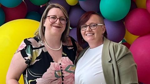 Zoe and Anna Lowey stand in front of a backdrop of colourful balloons. Zoe is wearing a floral top and has blonde hair and Anna is wearing a white top under a green jacket and has dark brown hair, both are wearing glasses.