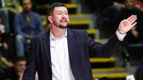 Marc Steutel, wearing a suit with an open-necked white shirt, makes a gesture with his left hand towards his basketball players