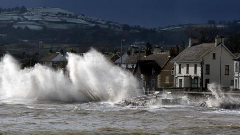 Waves crash against a promenade wall in a town on the Antrim coast in Northern Ireland. A row of detached houses is partially obscured by spray which is as high as the roofs. Behind the town are snow topped hills. 