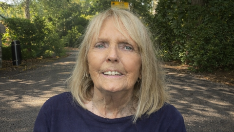 Jackie Whitehurst is looking at the camera and smiling. She has blonde, shoulder-length hair and is wearing a blue sweatshirt. She is in a park on a sunny day. 