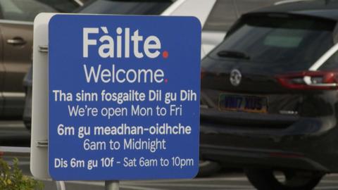 A blue sign with white writing outside Tesco in Stornoway. It reads "welcome" in English and Gaelic and includes information on opening times.