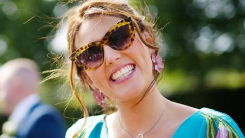 Mary has a beaming smile. Her light brown hair is blowing in the wind and she is wearing sunglasses, a colourful summer dress and pink flower earrings. 
