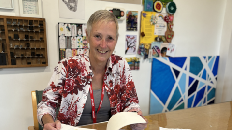 A woman in a red and white patterned top, sitting at a table with an open book and a collection of art work on the wall behind her