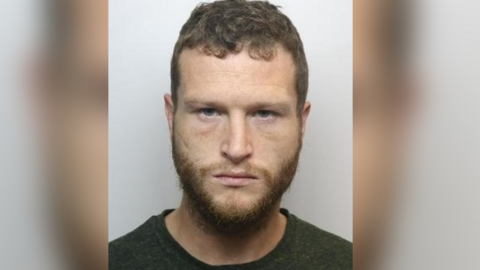 Levi Fishlock is shown in a police mug shot. He is staring directly at the camera, with short brown hair and beard. he is wearing a dark jumper with a round neck