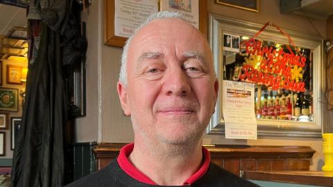 Man wearing a black jumper and red top sitting down in a pub with the beer taps and pictures in the background. 