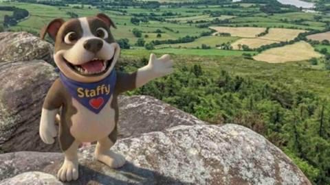 An animated design of a dog on its hind feet, waving with its paws towards the camera with a smile. The dog is brown and white with a blue neckerchief with "Staffy" and a heart on it. The dog stands on a rocky outcrop, overlooking green trees.