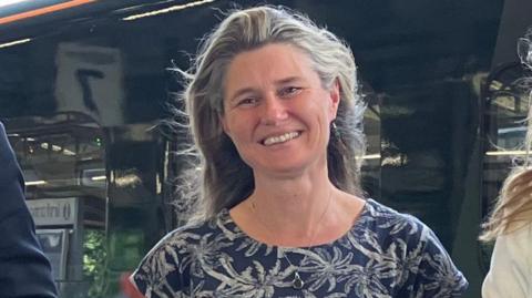 Jayne Kirkham - a woman with grey/dark hair smiling wearning a navy top with white tropical trees on it, background of a black train side