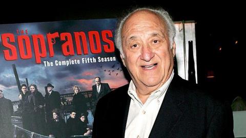 Jerry Adler, an elderly man, pictured at an evening event for The Sopranos TV series. He has short grey hair and wears a collared white shirt under a black jacket. He is stood in front of a poster for The Sopranos which reads: "The Sopranos: The complete fifth season" 